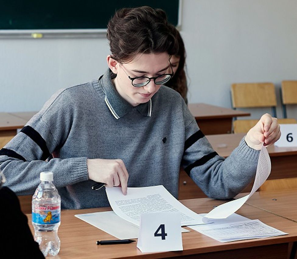 Kevin sitting at a desk looking at a test paper, wearing black glasses and a grey jumper - he has thick dark hair