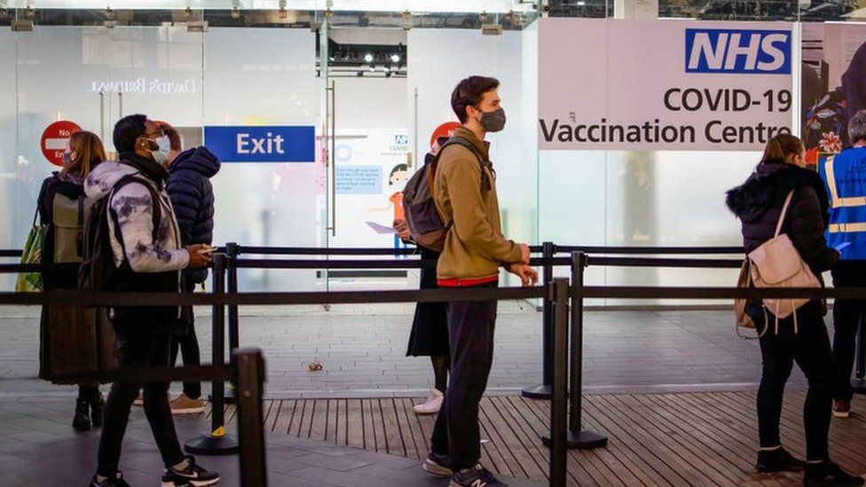 People are seen in a queue for their booster dose while standing at social distance outside the NHS vaccination centre at Westfield Stratford