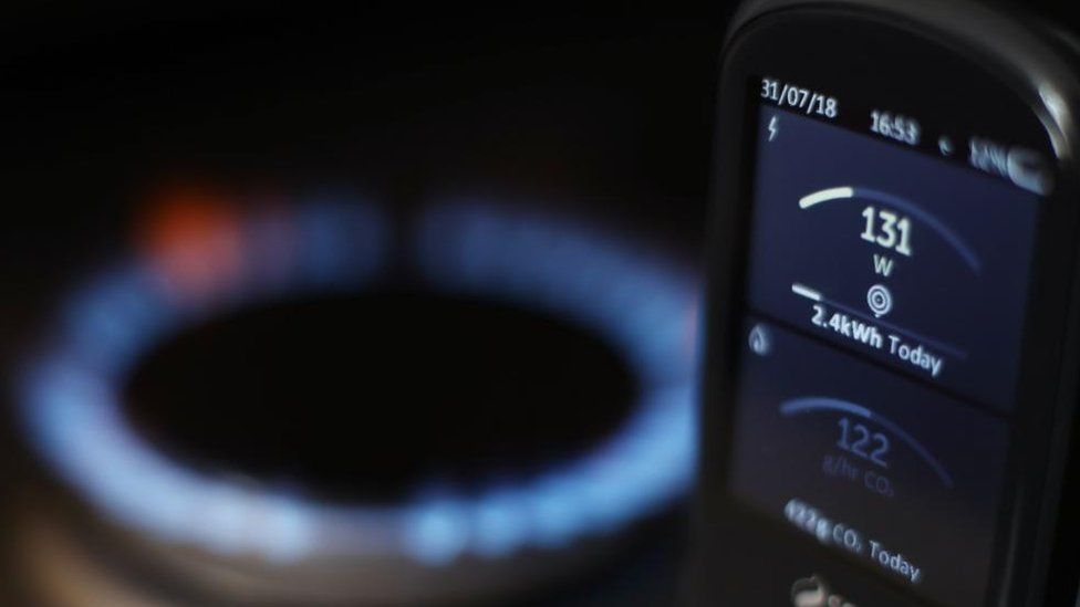 A smart meter showing energy usage. In the background, out of focus, is the flame of a burning hob.