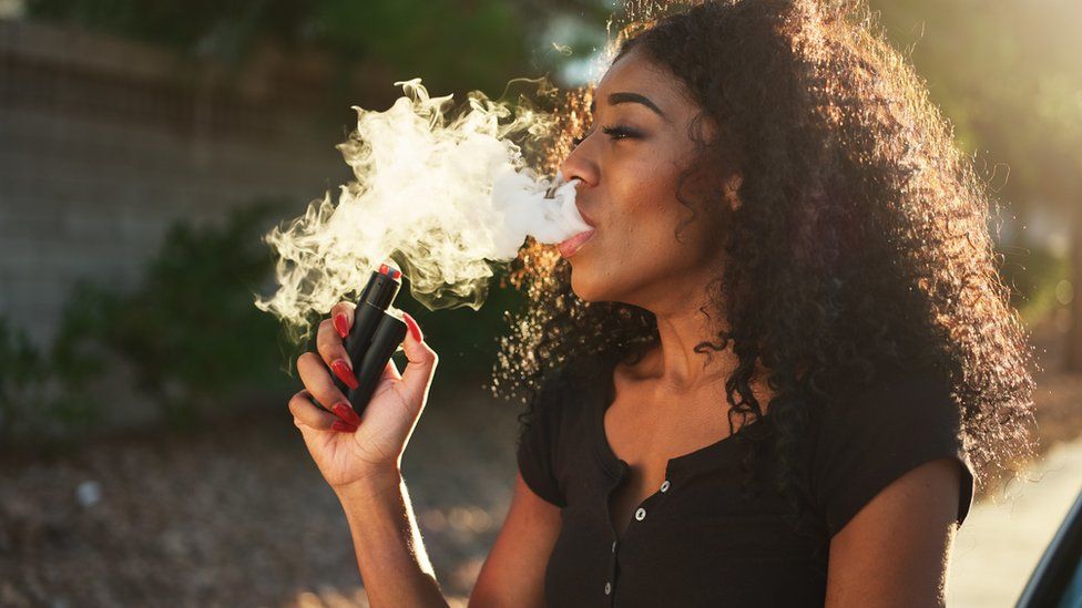 A woman, in a black t-shirt, exhales a large cloud of electronic cigarette vapour, as she is smoking from a vape. She has curly hair and is looking away from the camera
