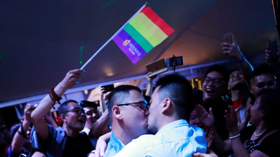 A gay couple kiss during a party on a cruise in open seas on route to Sasebo, Japan, 15 June 2017. A Pride flag hangs above them.
