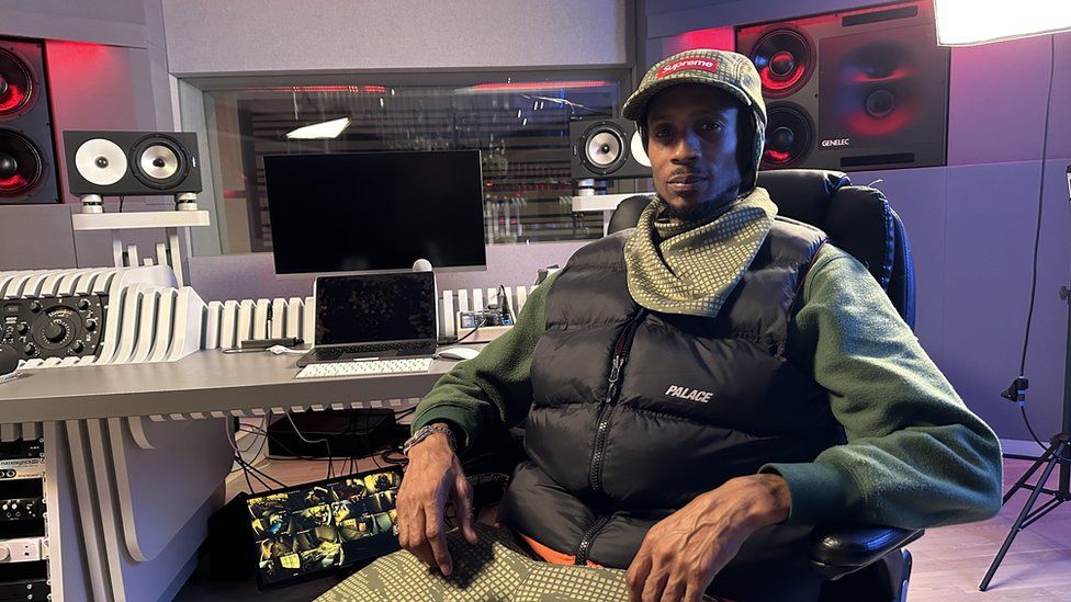 A man wearing a green hat and a green scarf with a black sleeveless jacket sits in a black chair in front of recording equipment in a music studio