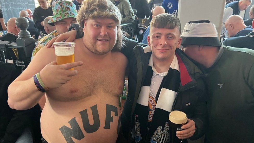 A topless man with NUFC tattooed across his belly stands next to a young man in a Newcastle shirt