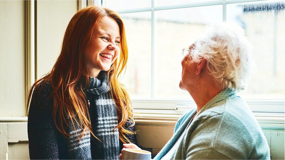 A granddaughter and her grandmother smile together beside a window