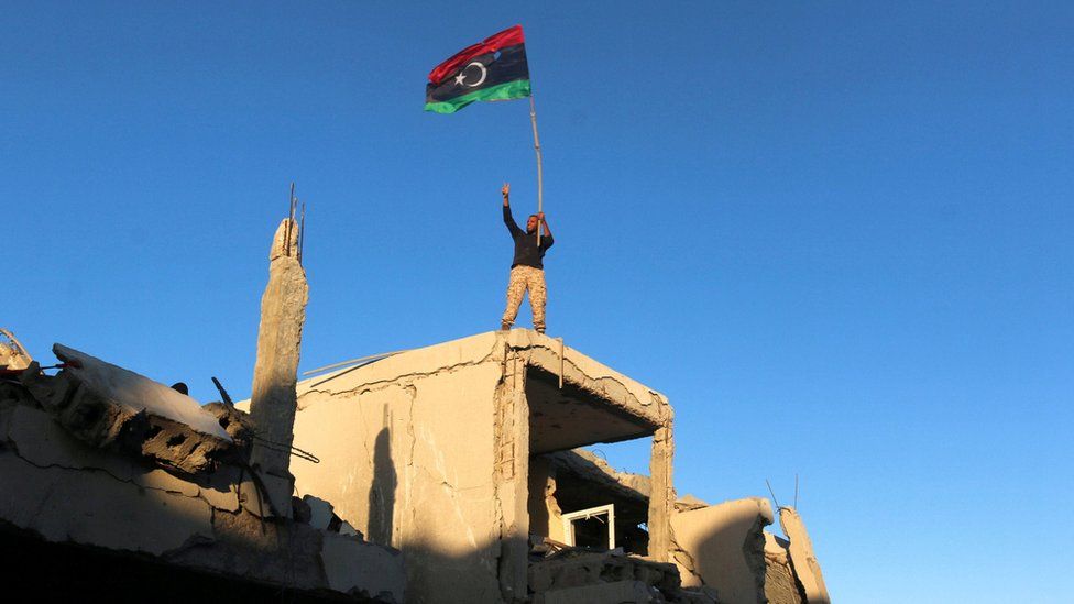 A fighter of Libyan forces allied with the U.N.-backed government waving a Libyan flag flashes victory sign as he stands atop the ruins of a house after forces finished clearing Ghiza Bahriya, the final district of the former Islamic State stronghold of Sirte, Libya