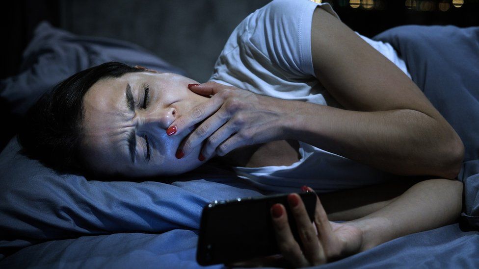 A woman yawns as she checks her smartphone while in bed