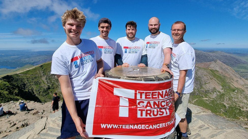 Luke, Elliot, and three others, holding a Teenage Cancer Trust banner at the summit of Snowdon