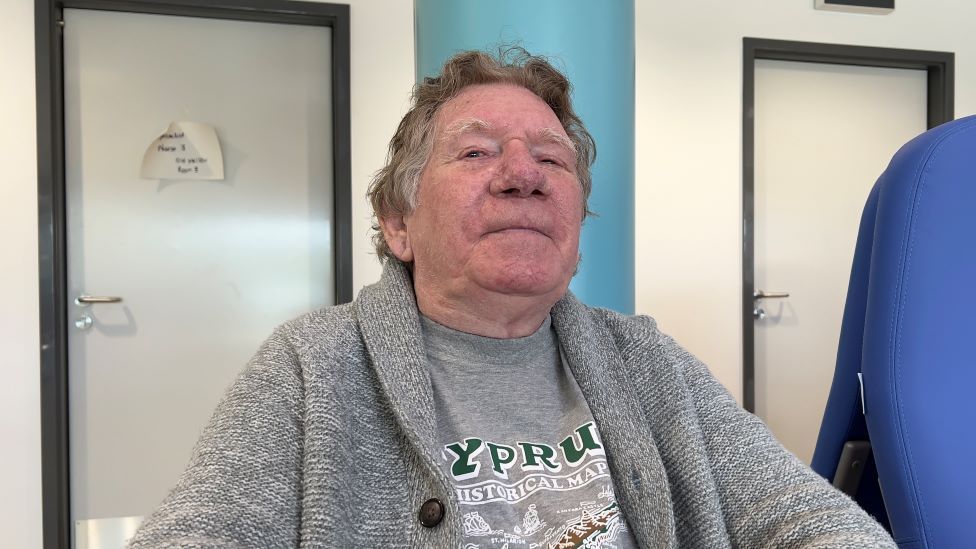 David Bancroft, de 76 años, está sentado con una camiseta gris y una rebeca mientras espera para ver a los especialistas de la columna vertebral en el Hospital de Ipswich