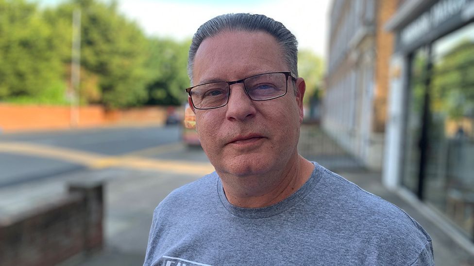 Head and shoulders picture of Carl Whitney, who has short, slicked-back, dark-grey hair and wears rectangular-shaped dark-framed glasses and a grey T-shirt.