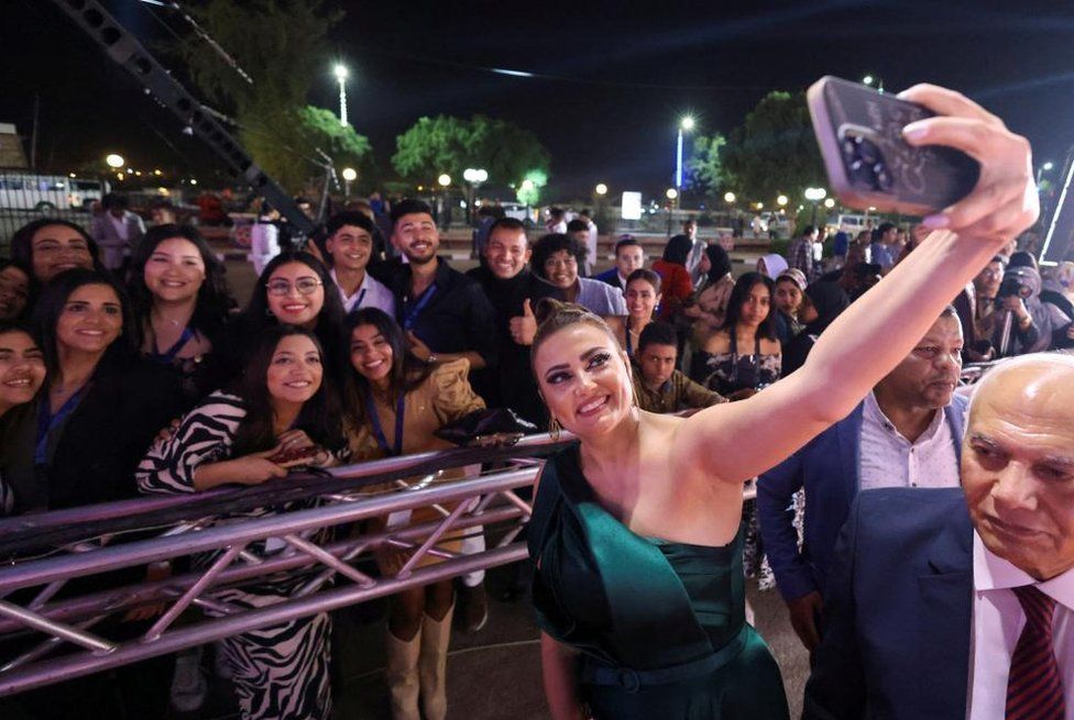 Actress Bushra holding up her phone posing for a selfie with several others in the background.