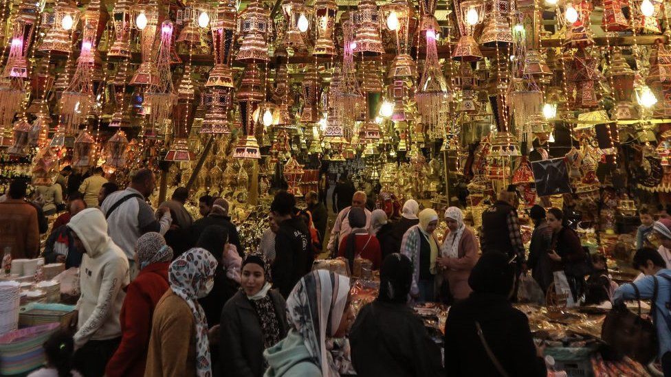 Women walk past a market stall with lit up lanterns. The stall area looks quite crowded.