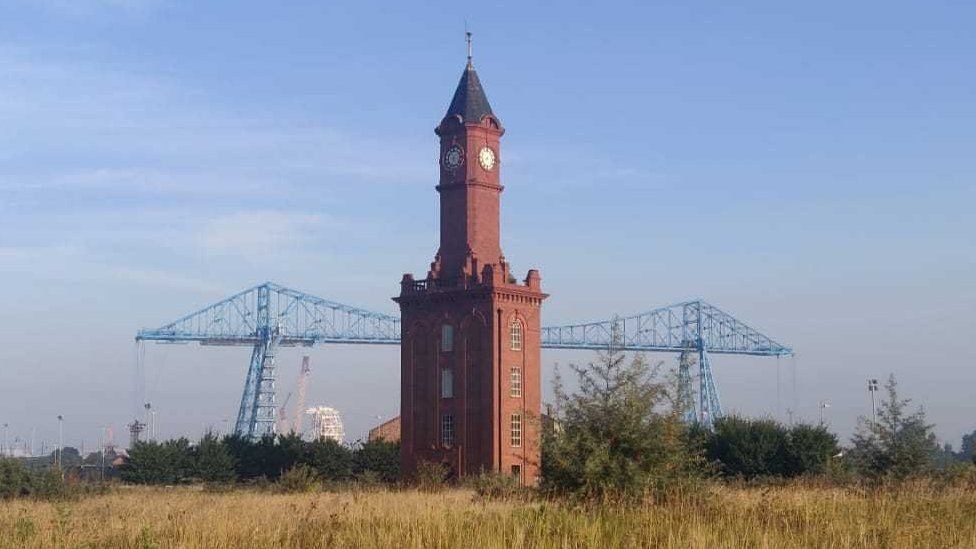 A red brick tower with clocks at the top with the blue transporter bridge beyond