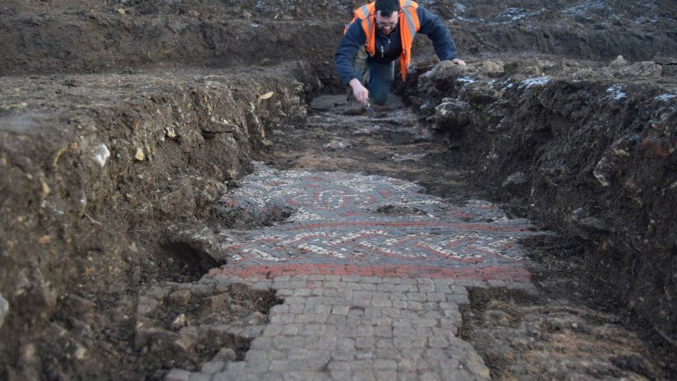 Photo at ground level shows part of the mosaic, including a red border, being carefully excavated by an archaeologist. The person is crouched over wearing a high visibility vest.
