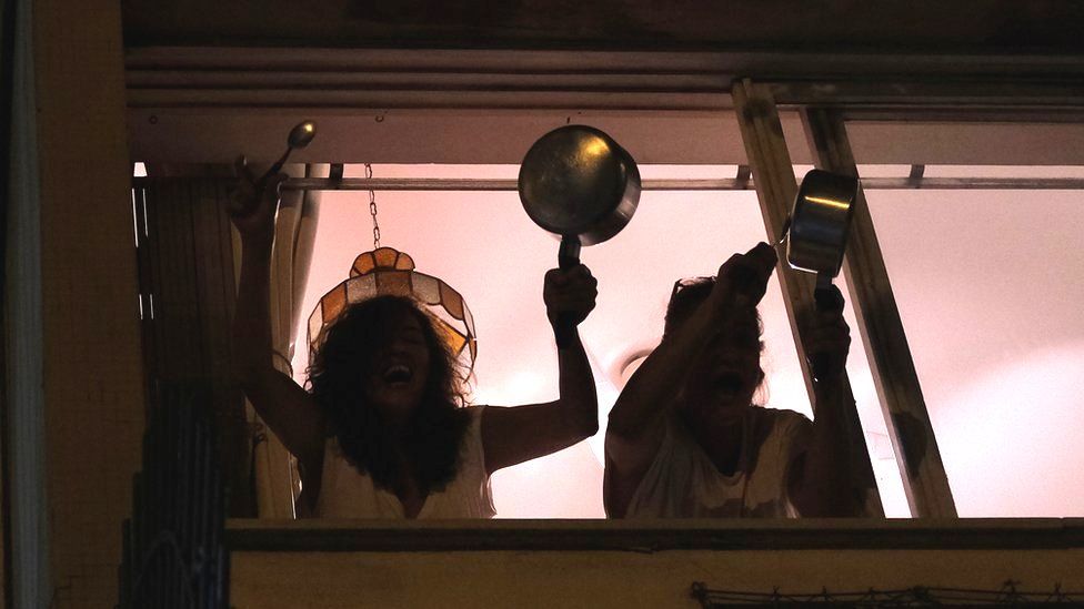 Women bang pots at the window of their apartment in Rio de Janeiro as they protest against Brazilian President Jair Bolsonaro over his handling of the coronavirus pandemic, 19 March 2020