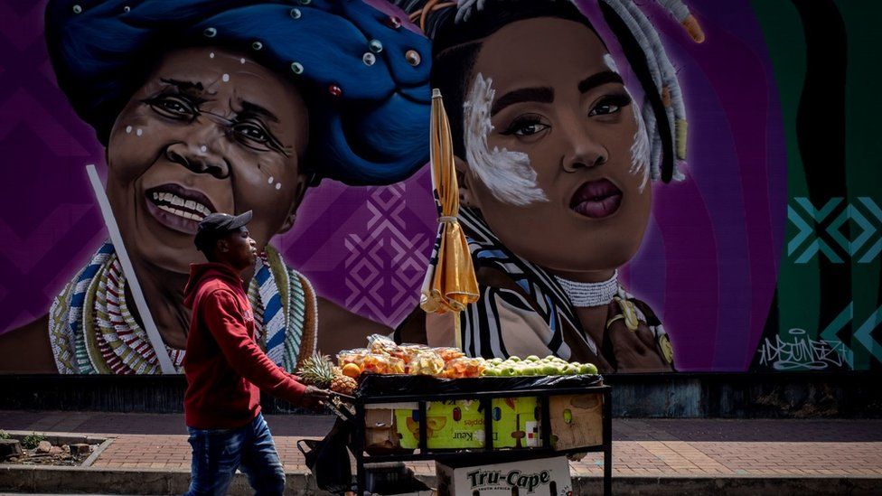 Man with a mobile fruit stall walking past a large street art mural. The mural shows two women. One is an elderly woman who is smiling. She has on a blue headdress and traditional neck adornments, the other lady is younger. She is pouting and has some white face paint on