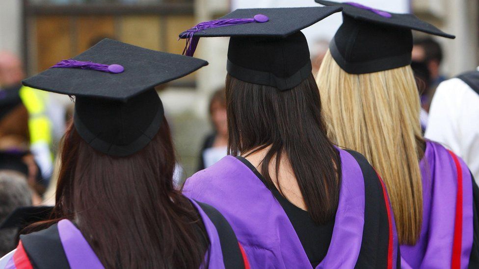 The back of three young women wearing black graduation gowns with purple and red detailing. They are also wearing a mortar board cap with purple tassels