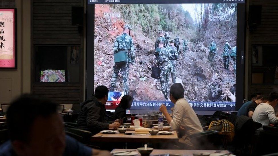 Посетители ресторана смотрят выпуск новостей по телевидению, в котором солдаты работают на месте крушения самолета в Учжоу