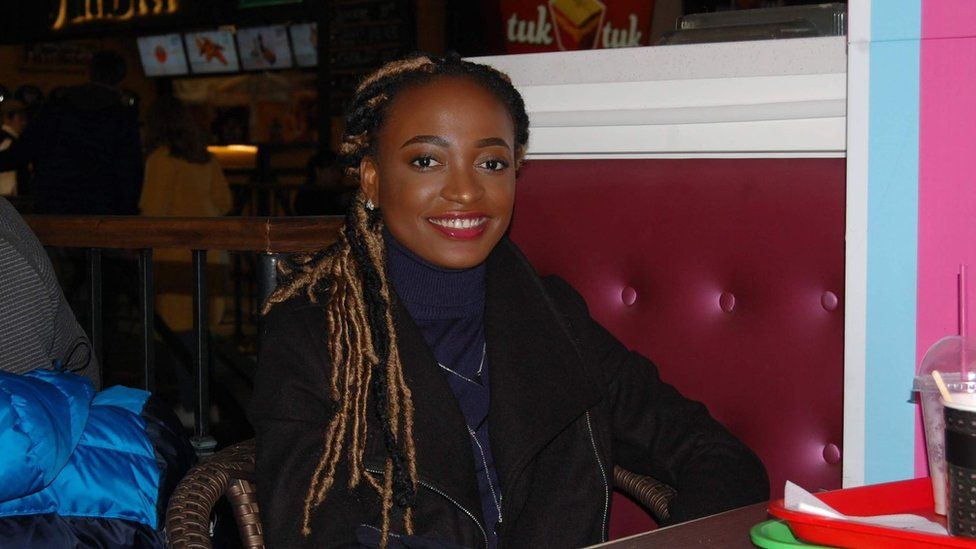 A woman with long hair styled in locs sits in a cafe or diner with red cushioned walls and signs advertising various food visible in the background. She's smiling and wearing a navy blue polo-necked top underneath a black jacket. She wears red lipstick and small, sparkly silver stud earrings.