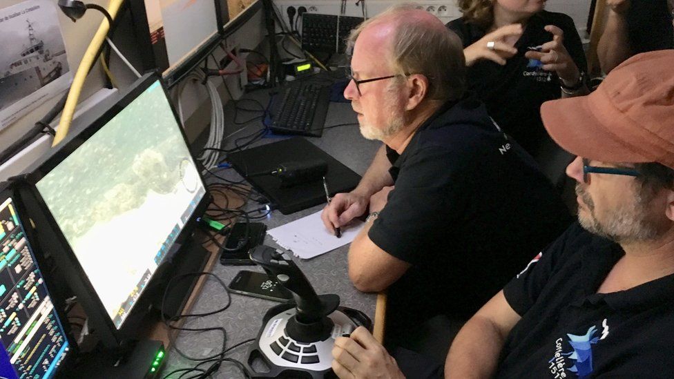 Crew members in casual clothing gather around a computer monitor, taking notes about the image on the screen - from our viewpoint, it is washed out and difficult to see the image