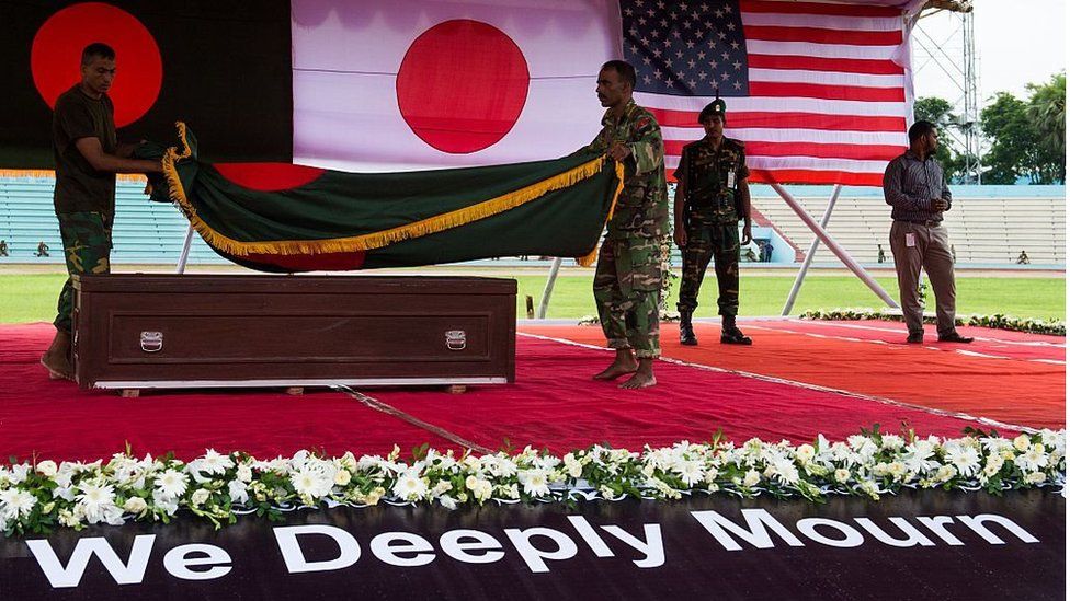 Бангладешские солдаты накидывают национальный флаг на гроб одной из бангладешских жертв теракта на фоне флагов Бангладеш, Японии и США во время поминальной службы в Дакке, 4 июля 2016 года.