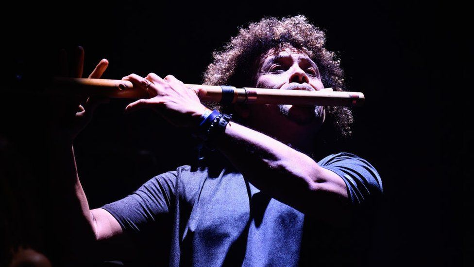 A man with curly, afro-style hair plays a modified woodwind instrument that resembles a flute. He's holding one end up to his mouth, and both arms are supporting the other end of the long instrument at chin height. He appears to be standing up, and exerting himself as he plays. He's bathed in a stage light that illuminates the front of his body and face but quickly fades off into blackness behind him.