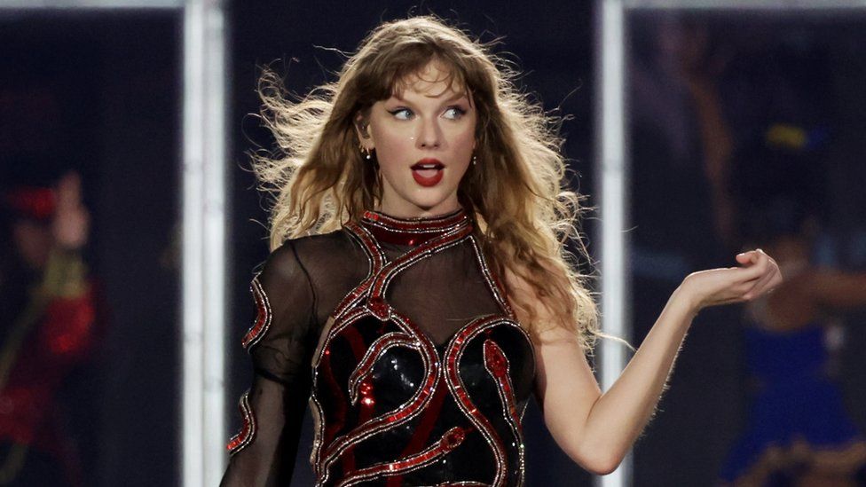 Taylor Swift performing in Singapore as part of her Eras tour. Taylor is a 34-year-old white woman with long wavy blonde hair and a fringe just above her eyebrows. She has blue eyes and looks to the left of the image. She wears her signature red lipstick and a black mesh one-sleeved jumpsuit which is embellished with red and black sequins and gems. She holds her right arm out, bent up at the elbow.