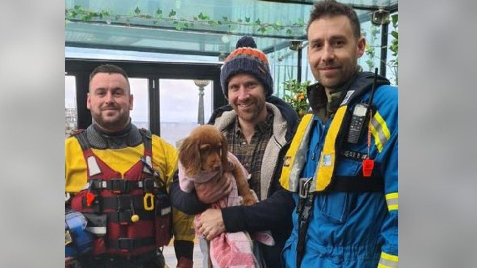 A man holding a ginger puppy next to two coastguard workers