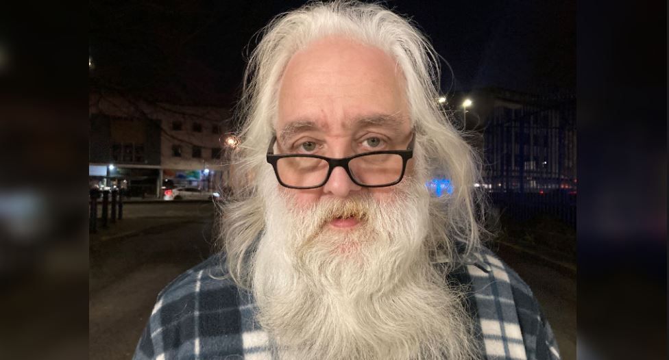 Jon Wisbey with long white hair and a beard, standing in a blue and white plaid shirt and wearing glasses