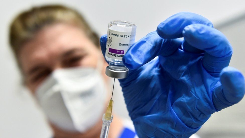 Someone taking a dose from an Oxford AstraZeneca bottle using a needle, holding the bottle up to the camera, with the bottle being held by a blue gloved hand