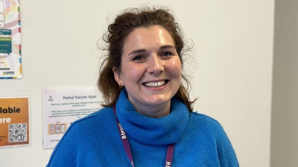 Nurse Clare Matthews at the Brook sexual health clinic in Bristol. Clare, a 31-year-old white woman, has curly brown hair down to her shoulders which she wears clipped back. She has brown eyes and small hooped earrings in her ears and is pictured smiling at the camera. She is wearing a royal blue turtle-necked jumper and a purple lanyard for work, and stands inside against a white wall with some posters on it for sexual health services.