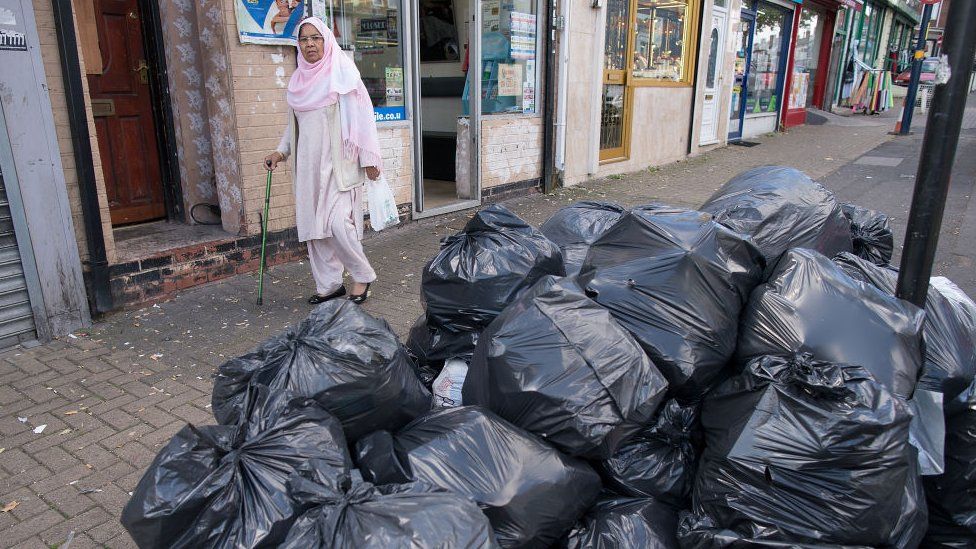 Woman walks past bags of rubbish in Alum Rock, Birmingham