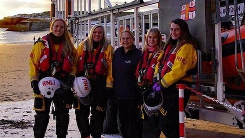 Five women stand next to a lifeboat