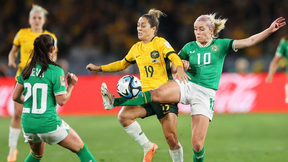 Australia football player Katrina Gorry and Republic of Ireland player Denise O'Sullivan competing for the ball during the Women's World Cup between the two sides. Denise is wearing a green Republic of Ireland kit with the number 10 on the front with white shorts and green socks. Katrina is wearing a gold Australia kit with green trim and the number 19 on the front. She is also wearing dark green shorts and white socks. Both players look like they are concentrating as they fight to get the ball which is closer to Denise's outstretched leg. There are also another Republic of Ireland and Australia player in the background of the picture to the left of Denise and Katrina.