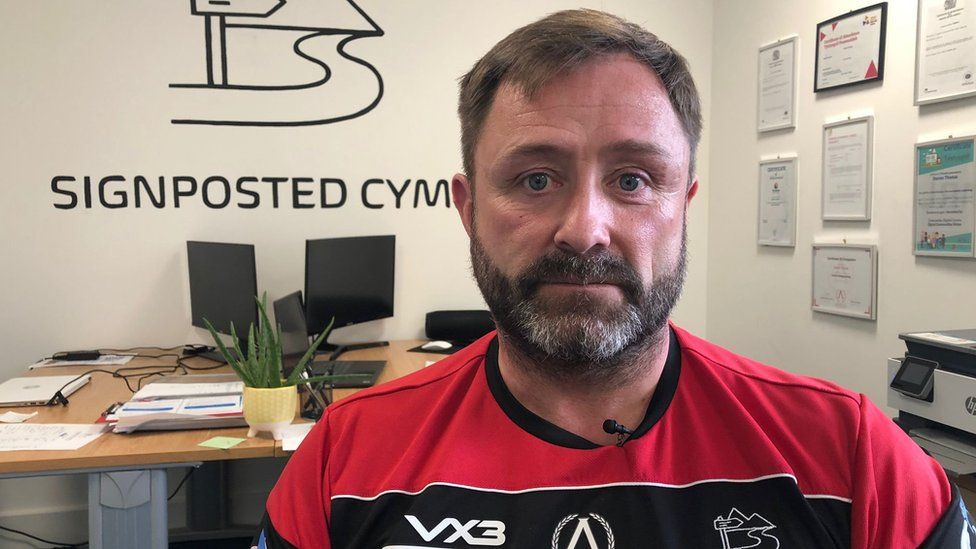 A man in a red and black sports top is looking at the camera. He has dark hair, with a beard and moustache. The sign on the wall behind him reads "Signposted Cymru".