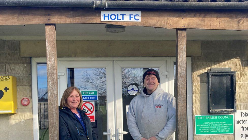 Nigel Tripp and Joy Bloomfield standing in front of the front doors with a small Holt FC sign above them