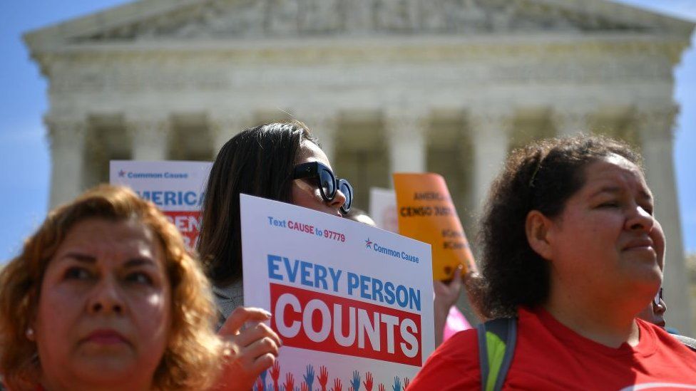 Three protestors outside the White House on 23 April. One holds up a sign saying "Every person counts".