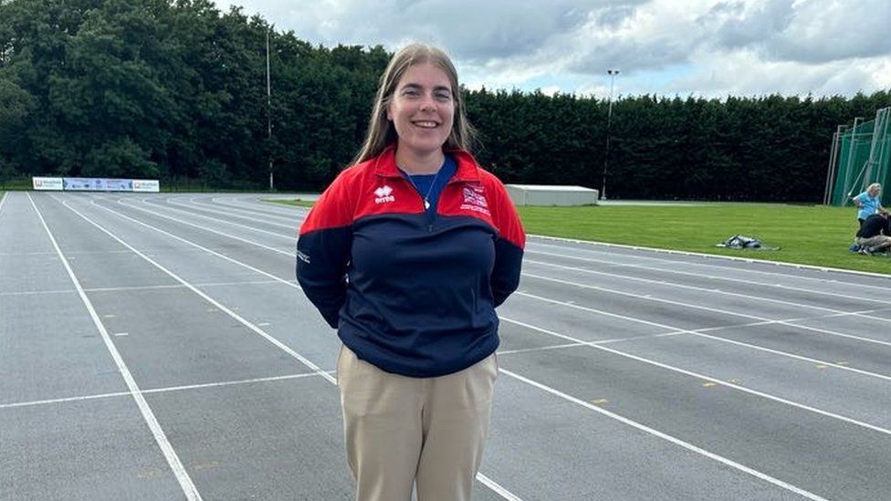 A woman in a blue and red Team GB jumper stands with her hands behind her back on an athletics track. She's got long, straight hair. The track is grey with white lines - the lanes curve off into the background and around a corner. A patch of grass where field events are held is also visible.