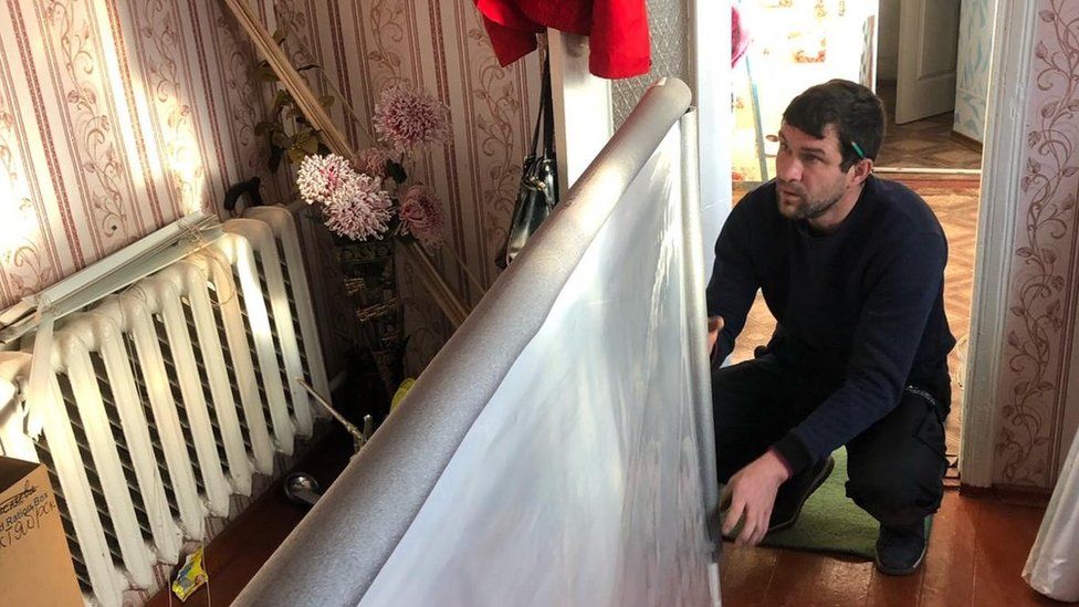 A man kneeling down holding a plastic window within a living room in Ukraine