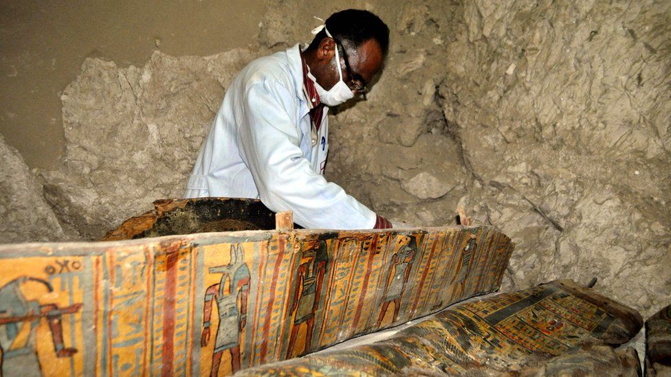 An archaeologist in a mask working on a colourfully-painted coffin inside a tomb