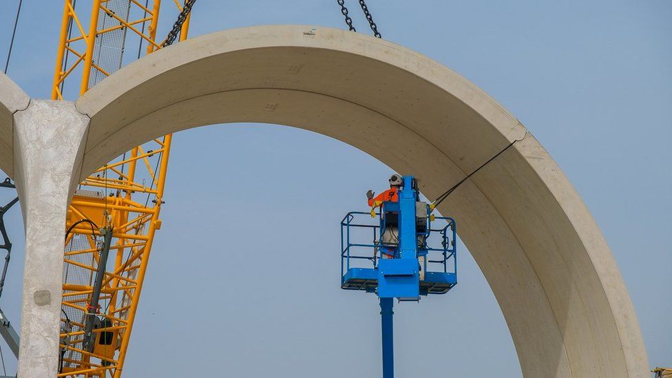 Crane with man on platform lifting concrete arch into place