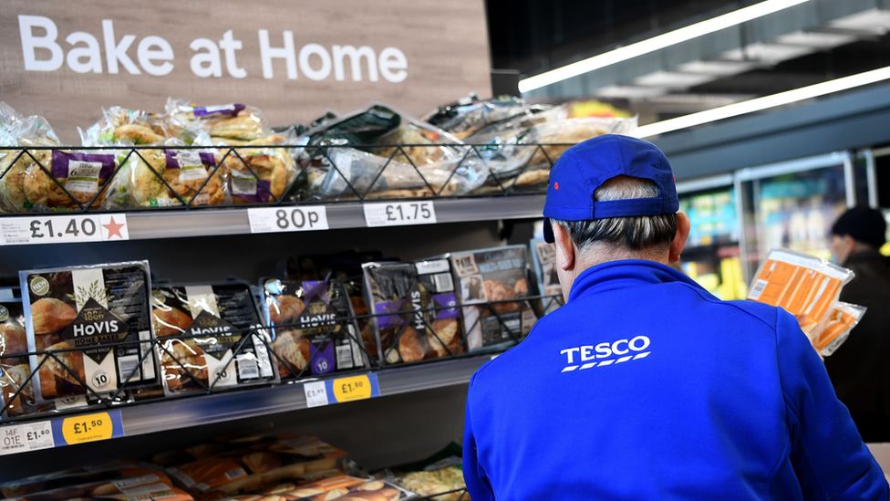 A Tesco employees stands with their back to a set of shelves showing different types of bread for sale in the shop. The employee is wearing a blue hat and fleece, both of which have Tesco branding.