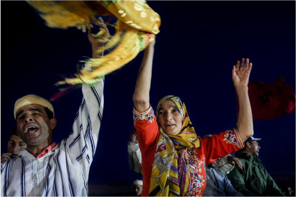 Woman wearing a yellow hijab and red outfit waving a yellow scarf in the air. She is next to a man who looks as though he is singing or cheering. The background is pitch black.
