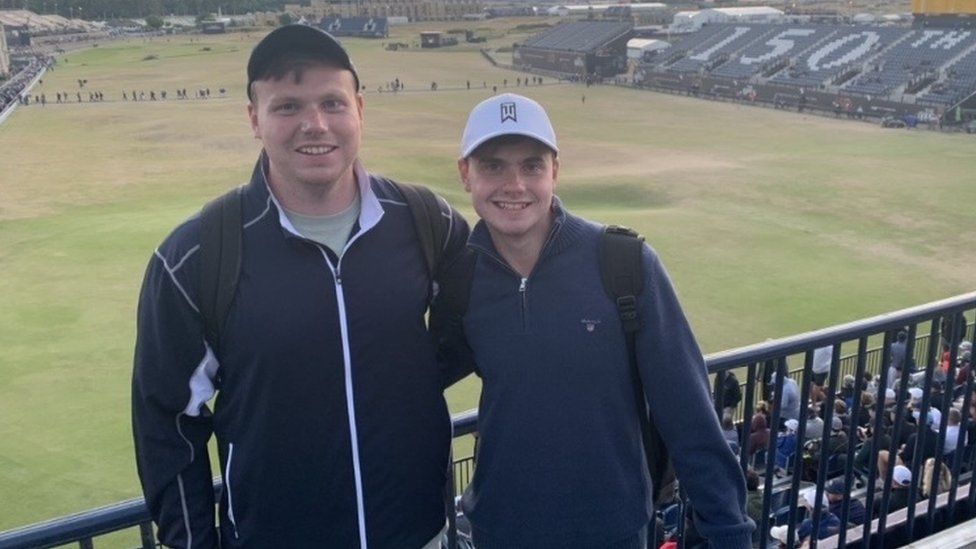 Ben and Jack O'Sullivan pictured at a sports event. Ben, left, is a 27-year-old white man and has his arm around Jack, right, who is slightly shorter than his older brother. They are both wearing caps - Ben's is dark and Jack's white, and dark zip-up jumpers. They stand against a railing which separates them from other spectators who are looking ahead at a wide grassy area,