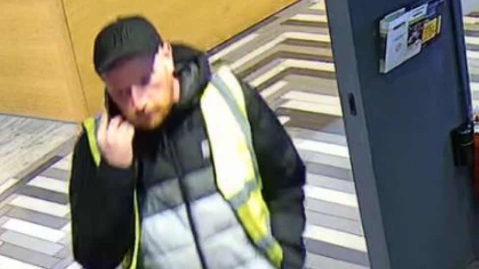 CCTV image of a man whit facial hair. He is wearing a black cap, a black, white and grey jacket partially covered by a yellow high viz jacket