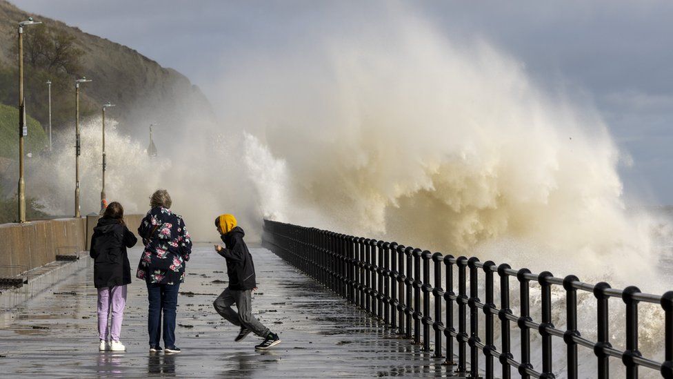 Image of people walking along the sea defence promenade walkway while waves crash over them on Sunny Sands Beach, Folkestone
