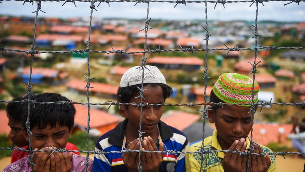 Image of children praying behind a barbed-wire fence. In February of this year, the Bangladeshi authorities began erecting barbed-wire fences around the camps, they say, "to prevent criminal activities"