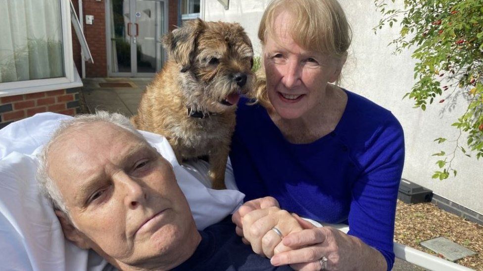 Tony Hughes lying outdoors in a hospital bed with his wife Sue and a small dog