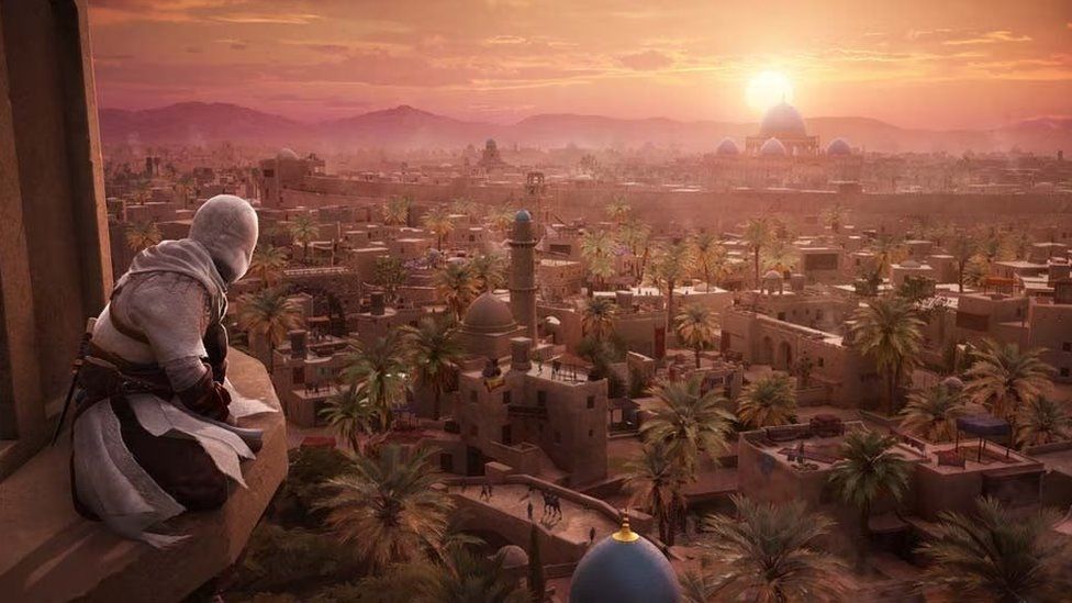 A scene from Assassin's Creed Mirage shows a figure dressed in white, his head covered with a hood, perched on a high ledge on a tall building. He's looking out over 9th Century Baghdad - a sprawling town in the desert. The sun creates a purple glow over the city as it sets, lighting up the collection of flat-topped buildings and domed structures that were typical of the period. The image suggests a vast city to explore