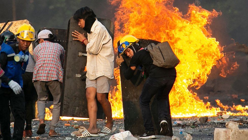 Протестующие за щитами на горящей улице в Мьянме.
