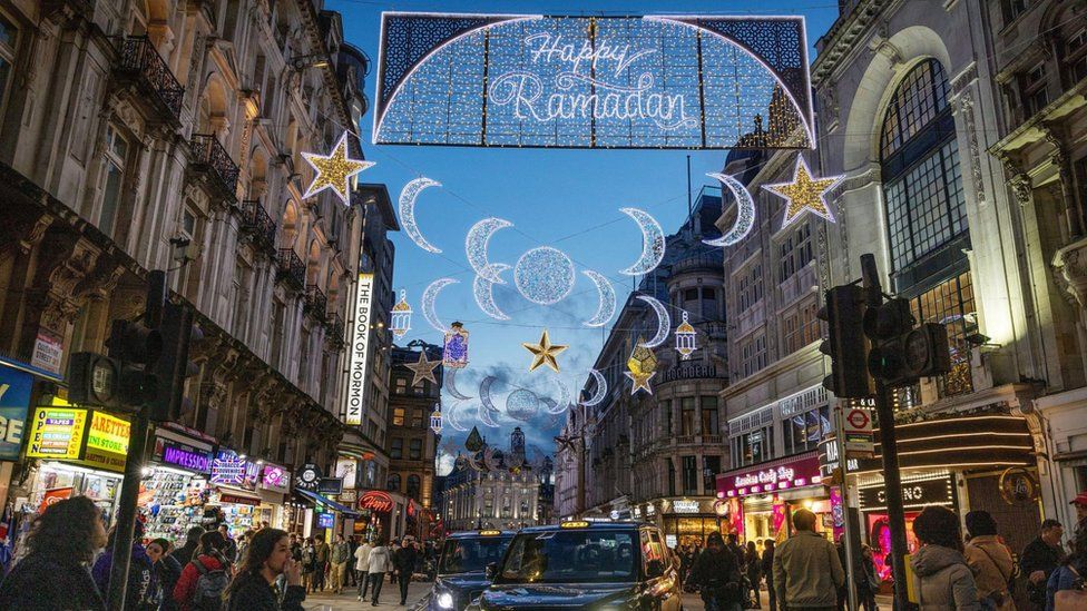 Happy Ramadan lit up in lights in Piccadilly as people look on and a taxi drives past